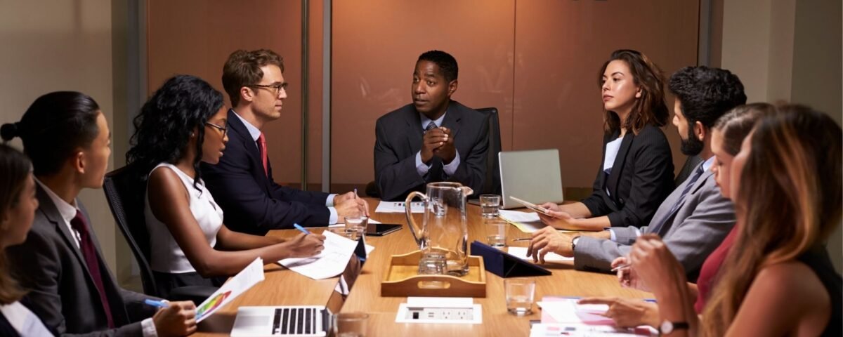 Group of professionals in a boardroom seated around a wooden table, with a man speaking in the center as others listen and take notes.