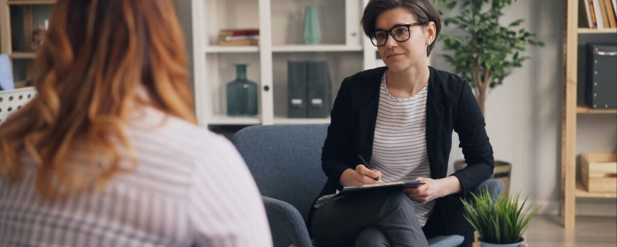 Therapist with short dark hair and glasses sits with a clipboard, listening to a client in a bright, plant-filled office.