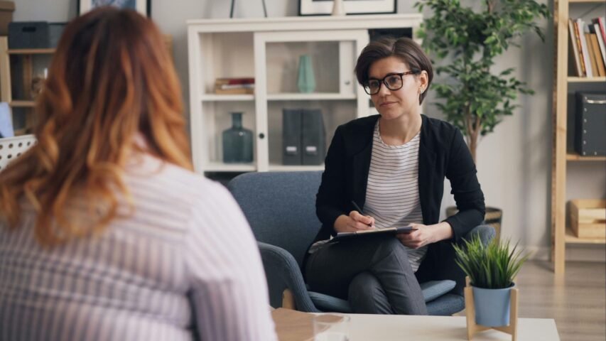 Therapist with short dark hair and glasses sits with a clipboard, listening to a client in a bright, plant-filled office.